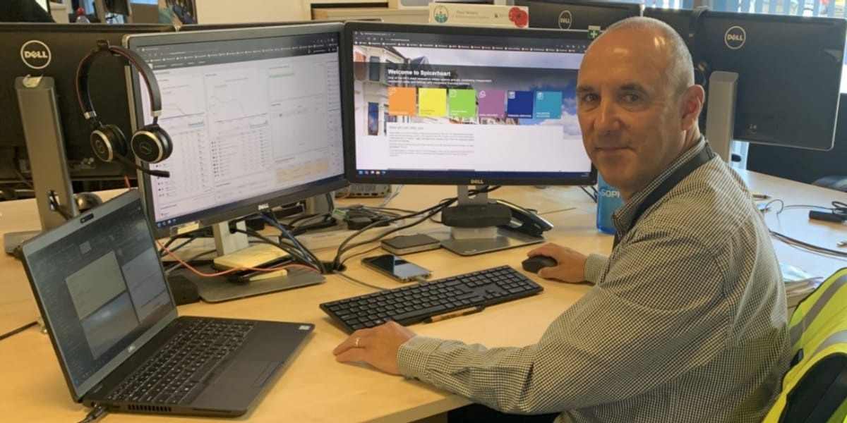 A person sits at a desk with two monitors, a laptop, and headphones, engaged in work within an office environment.