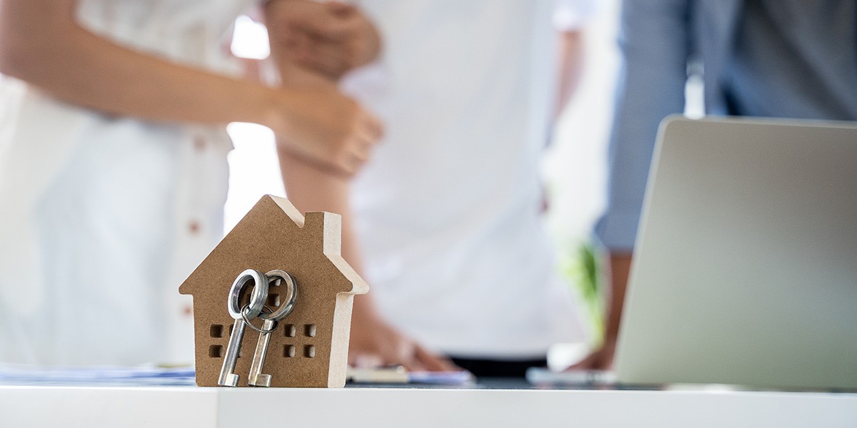 A decorative wooden house with keys in front of a couple discussing at a desk, symbolising home ownership and new beginnings.