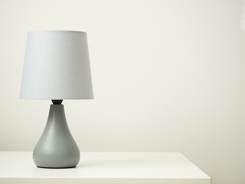 A modern lamp with a round, striped wooden base and a cylindrical white shade sits on a sleek white shelf against a light wall.